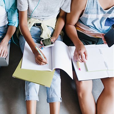 Close-up overhead portrait of girl in blue shirt and jeans holding laptop on knees while sitting beside university mates. Female student writing lecture in notebook and using phone between friends.. Close-up overhead portrait of girl in blue shirt and jeans holding laptop on knees while sitting beside university mates. Female student writing lecture in notebook and using phone between friends..