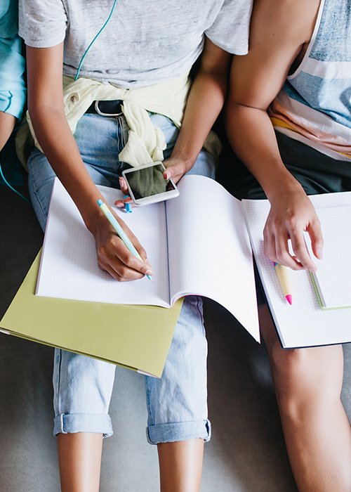 Close-up overhead portrait of girl in blue shirt and jeans holding laptop on knees while sitting beside university mates. Female student writing lecture in notebook and using phone between friends..