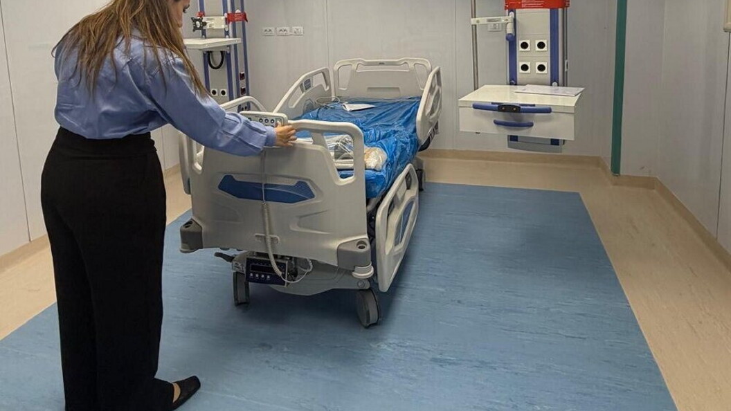 A nurse adjusts a hospital bed in a ward with blue flooring