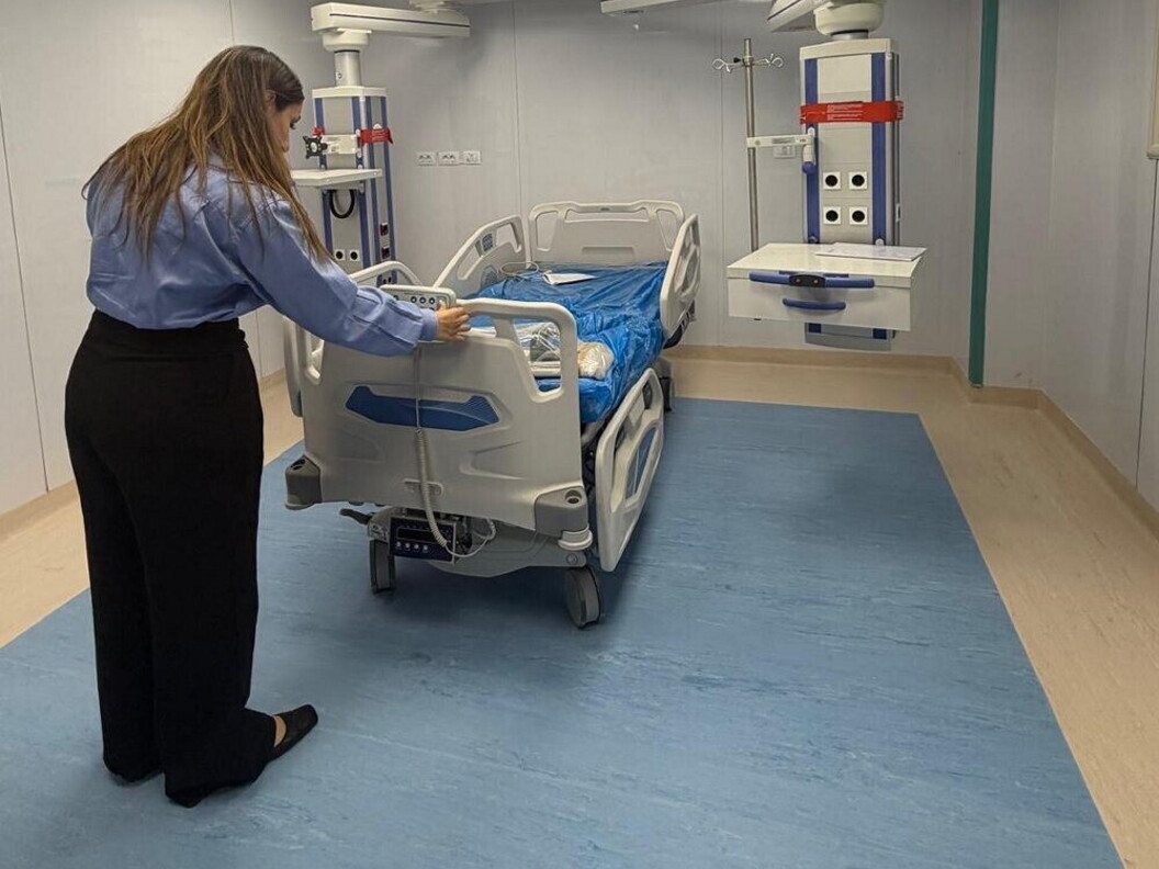 A nurse adjusts a hospital bed in a ward with blue flooring